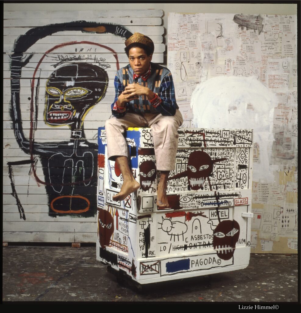 Jean-Michel Basquiat retratado en su estudio, sentado sobre una caja pintada, ante dos de sus obras