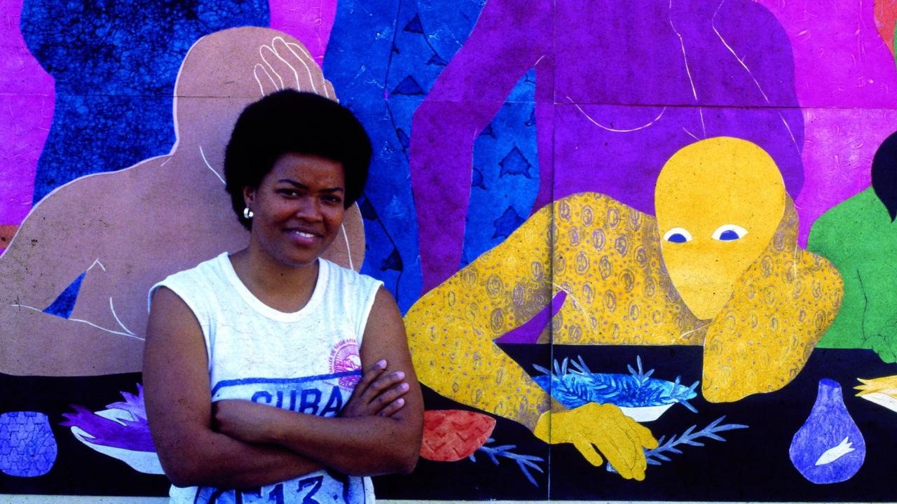 Belkis Ayón, joven, con los brazos cruzados y sonriendo, ante uno de sus raros murales en color con figuras azules, moradas y amarillas