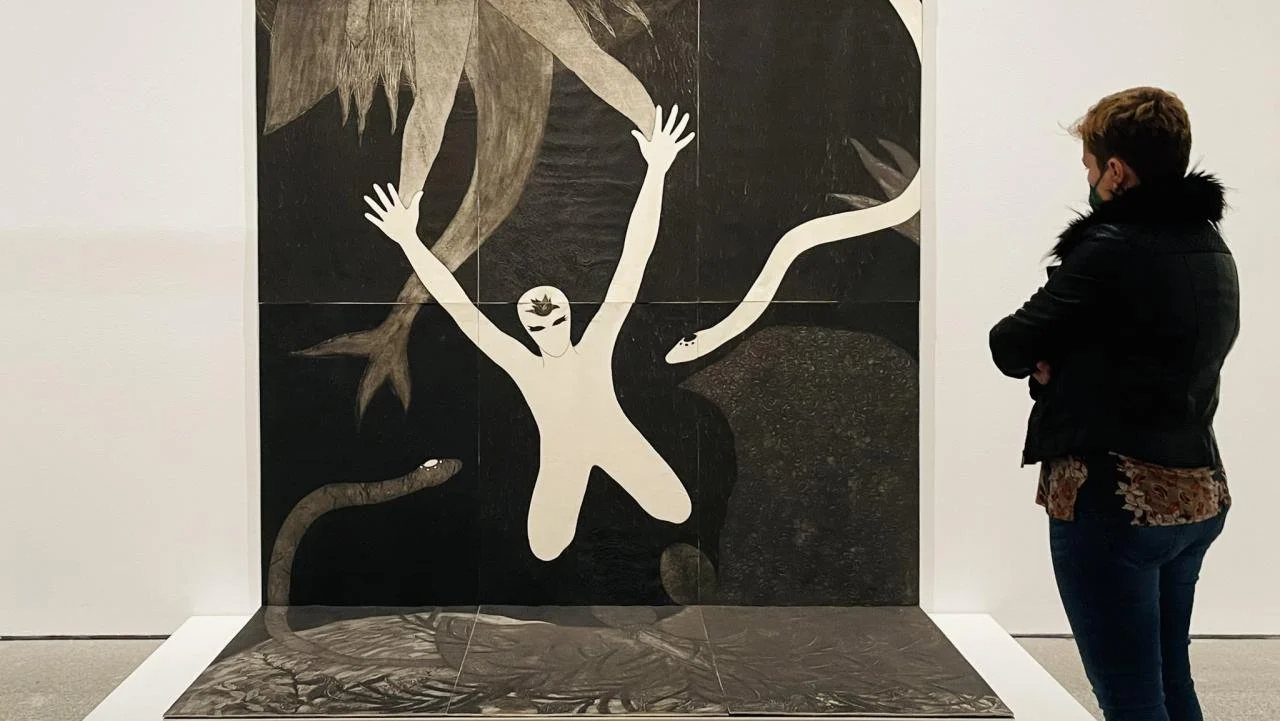 Visitante ante una gran colografía de Belkis Ayón con una figura blanca de brazos en alto entre serpientes, en una sala de museo