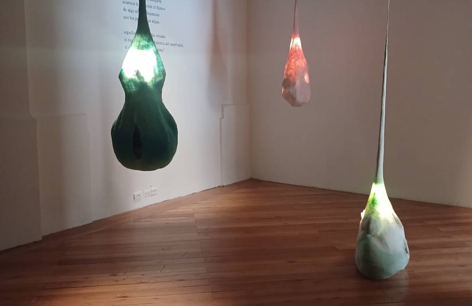 Tres esculturas lumínicas en forma de gota, en tonos verde y rojo, suspendidas y posadas en el suelo de una sala junto a un poema proyectado en la pared
