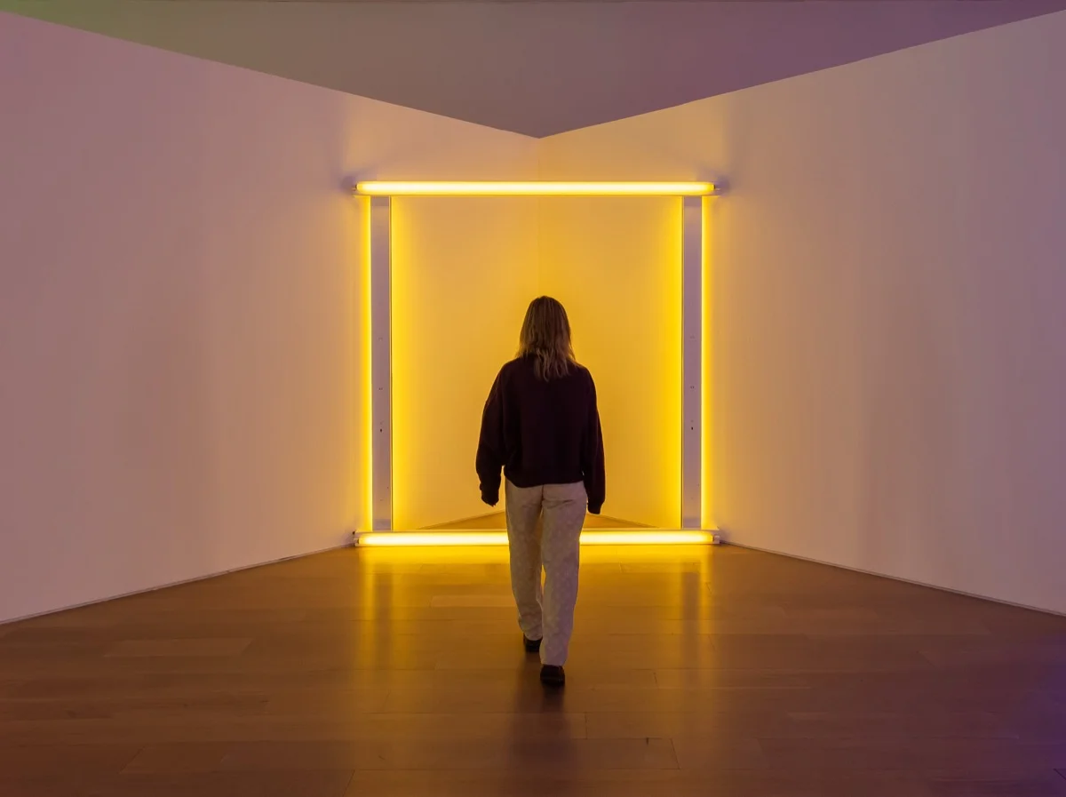 Instalación de Dan Flavin: un cuadrado de tubos fluorescentes amarillos que inunda de luz dorada una sala blanca; una figura de espaldas camina hacia el resplandor